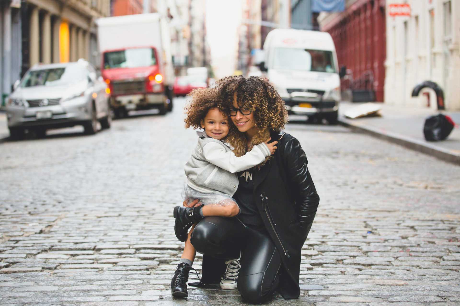 young mother holding a baby on a cobblestone street with commercial trucks in the background