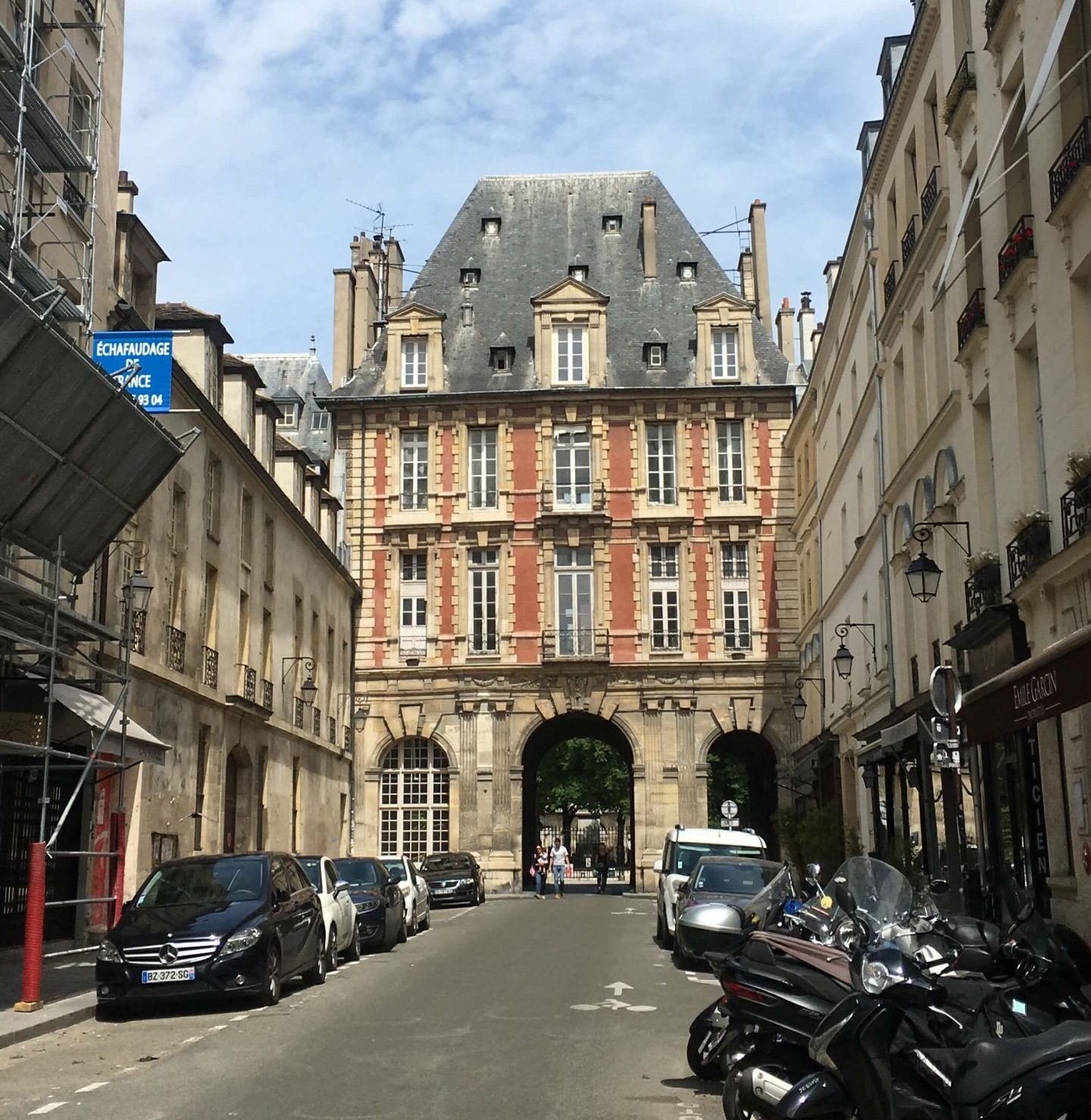 Street lined with cars and scooters leading up to brick and stone building