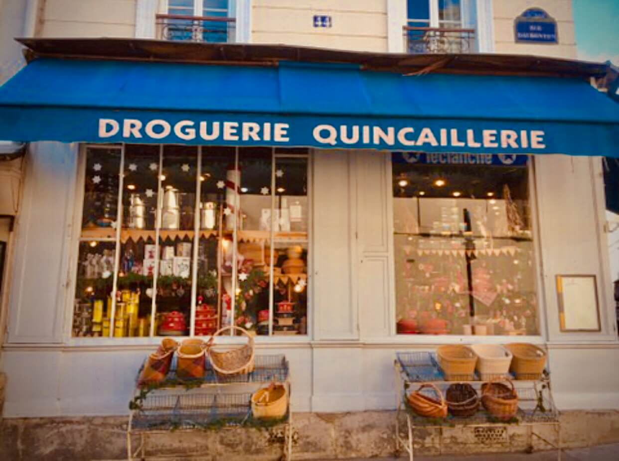 Blue awning over two windows says Droguerie Quincaillerie. Baskets sit on shelves on the sidewalk