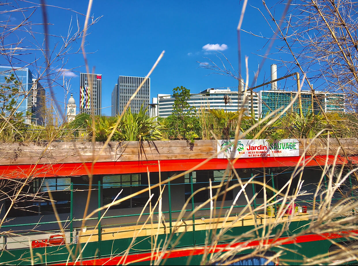 A boat named Jardin Sauvage with many green plants and the city skyline behind it with a bright blue sky