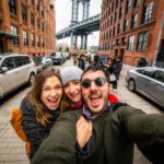 Friends taking selfie in front of Manhattan Bridge as seen from Dumbo, Brooklyn, New York City-min