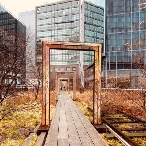 Walk along the High Line in New York City, passing through a series of rectangular metal frames on a wooden pathway, surrounded by winter plants and modern glass buildings.