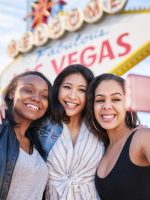 three friends on vacation taking a group selfie in front of the welcome to las vegas sign