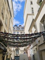 Decorative string lights and classic Parisian balconies over a narrow Le Marais street.
