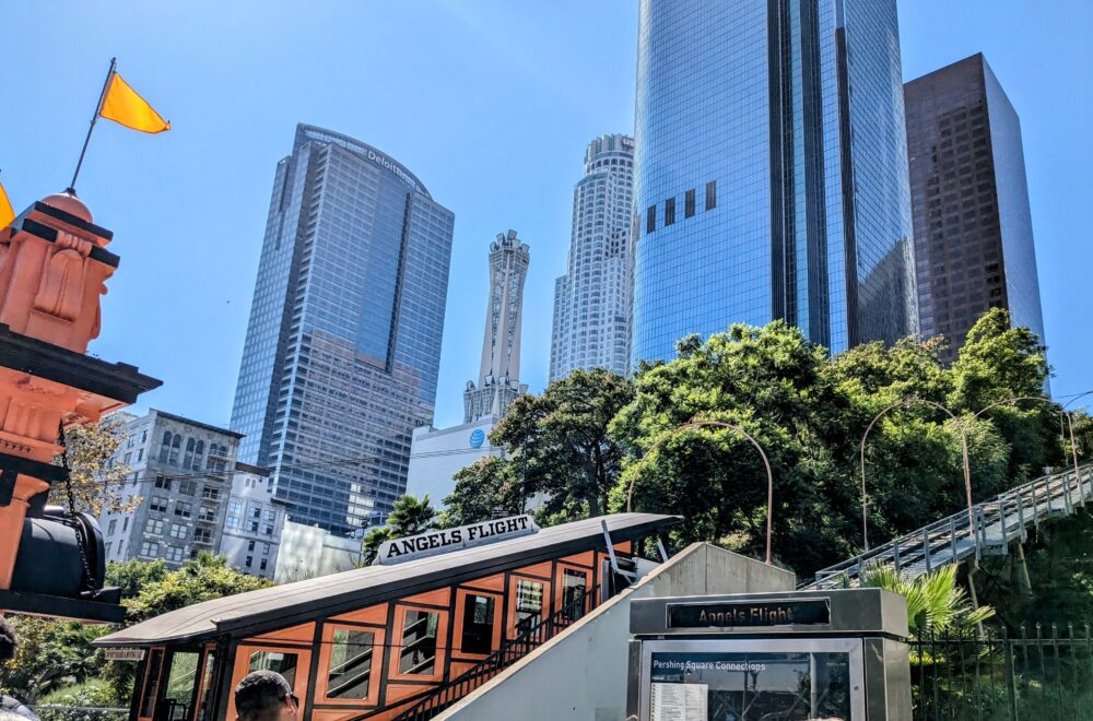 Tourists taking photos near Angels Flight Railway with downtown LA skyscrapers in the background on a sunny day.