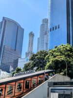 Tourists taking photos near Angels Flight Railway with downtown LA skyscrapers in the background on a sunny day.