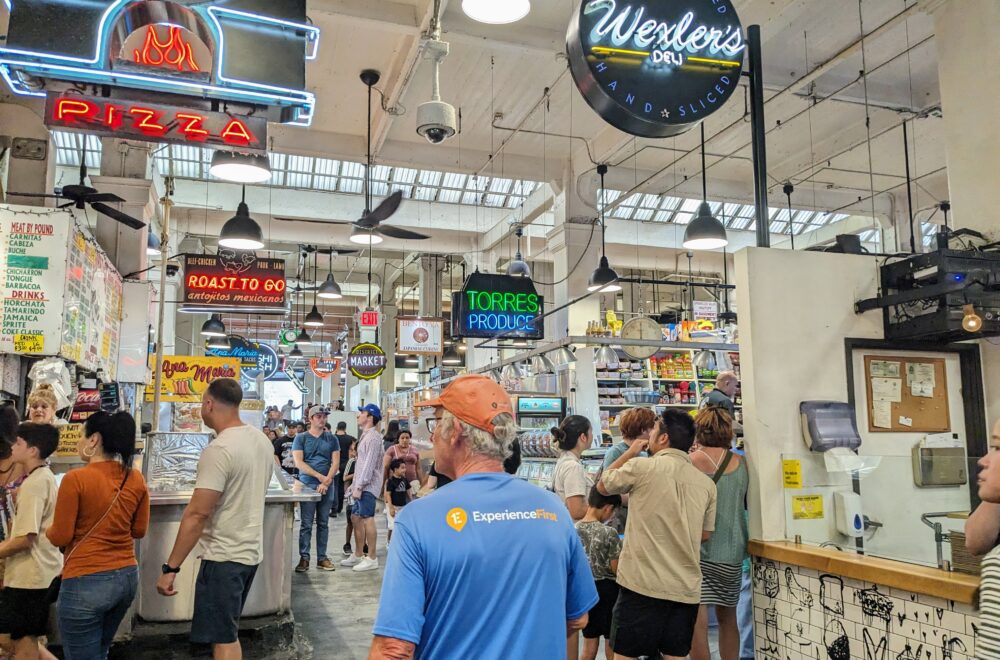 Visitors exploring food stalls at Grand Central Market in Los Angeles, with hanging neon signs and a lively crowd.
